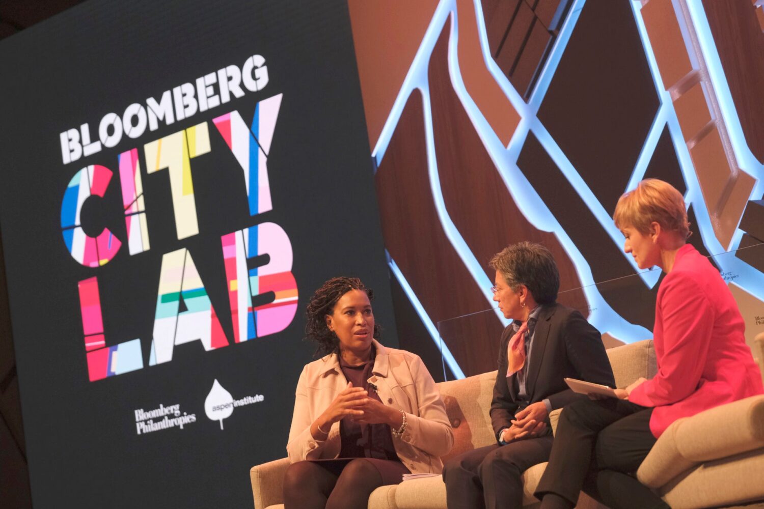 Three women sit on a stage for a panel, with a large colorful sign that says "City Lab" and the Bloomberg and Aspen Institute logos behind them. The woman on the far left, DC Mayor Muriel Bowser, is black with short hair and is wearing a tan button down. She is speaking and gesturing with her hands. To her right are two other women. The middle one has short salt and pepper hair, glasses, a black blazer and a neckerchief on. To the far right is a woman with short blonde hair and a pink jacket. Both are listening to Mayor Bowser speak.