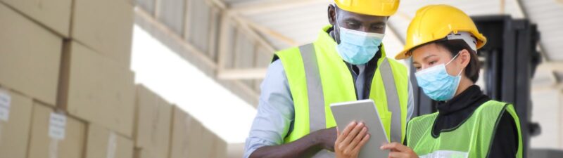 Photo of two warehouse workers wearing hard hats, reflector vests, and face masks. They are standing in front of a stack of cardboard boxes while looking at an electronic tablet. One is a Black man and the other is an Asian woman.