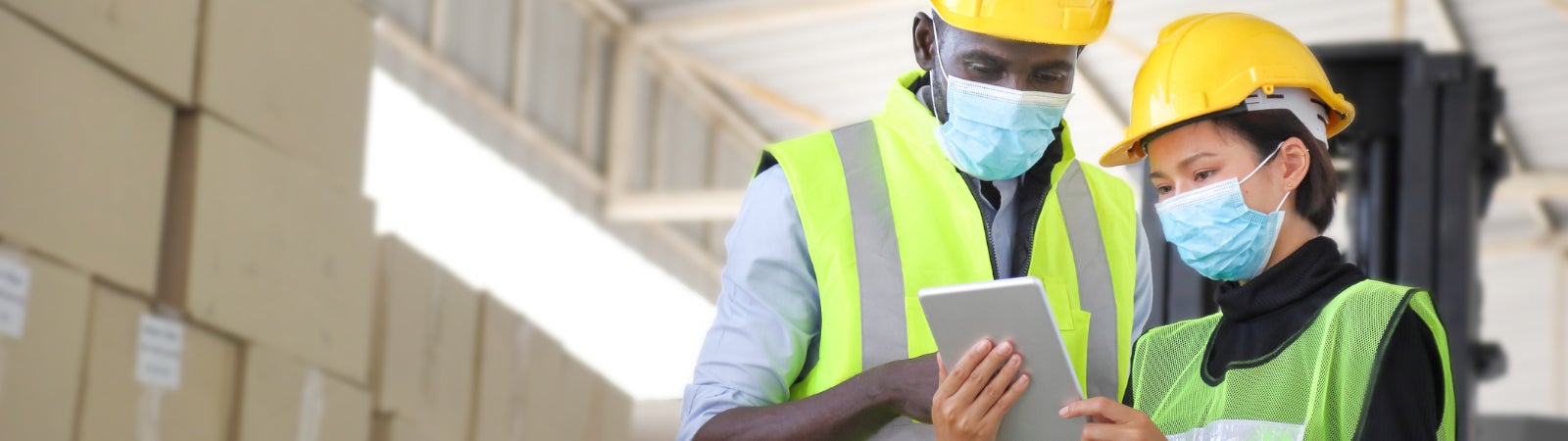 Photo of two warehouse workers wearing hard hats, reflector vests, and face masks. They are standing in front of a stack of cardboard boxes while looking at an electronic tablet. One is a Black man and the other is an Asian woman.