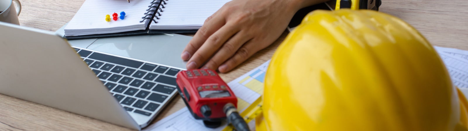 A photo of a man working at a desk with a hard hat, a laptop, and a handheld radio in front of him