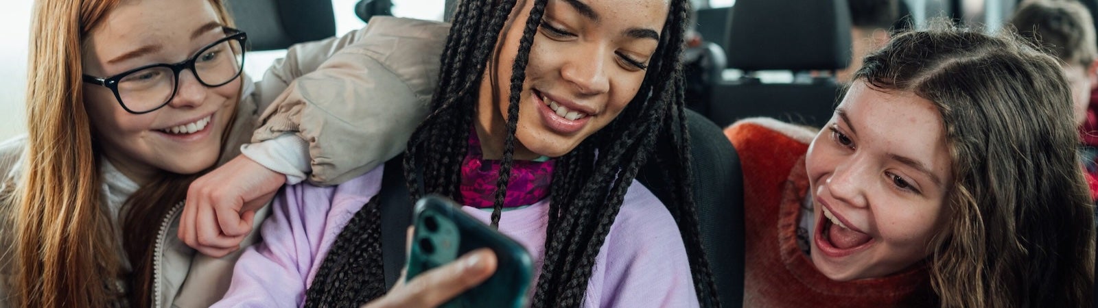 A photo of a group of three teenagers on the bus. They are looking a mobile phone and smiling and pointing at the screen.