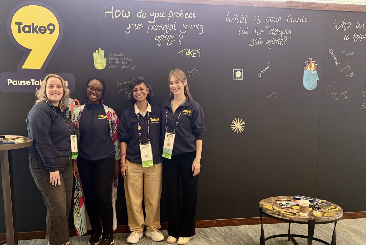 A photo of the Aspen Digital team with CyberFareedah in front of the Take9 chalk wall at WiCyS Conference.
